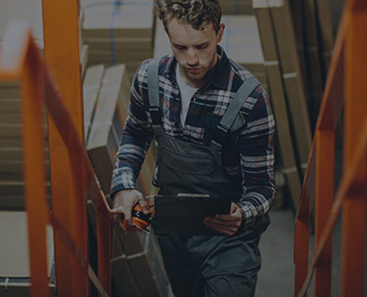 A worker in overalls holding a tool and a phone on stairs.