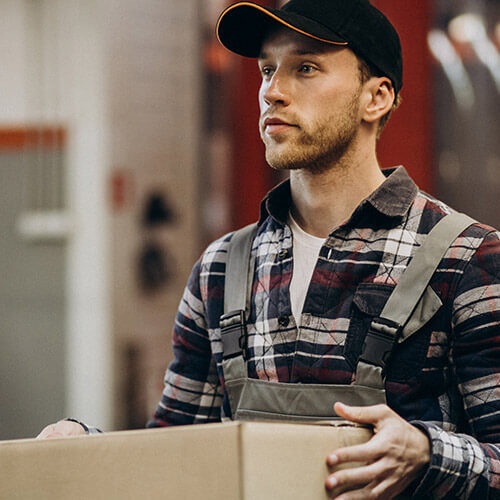 Man in cap and checkered shirt carrying cardboard boxes indoors.
