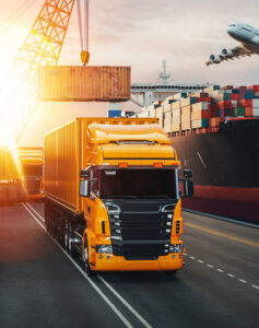 A yellow cargo truck drives near shipping containers at sunset.