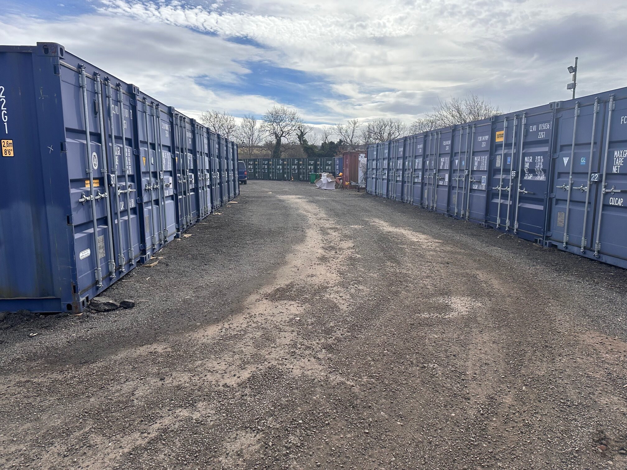 A gravel path between rows of blue shipping containers under a cloudy sky.